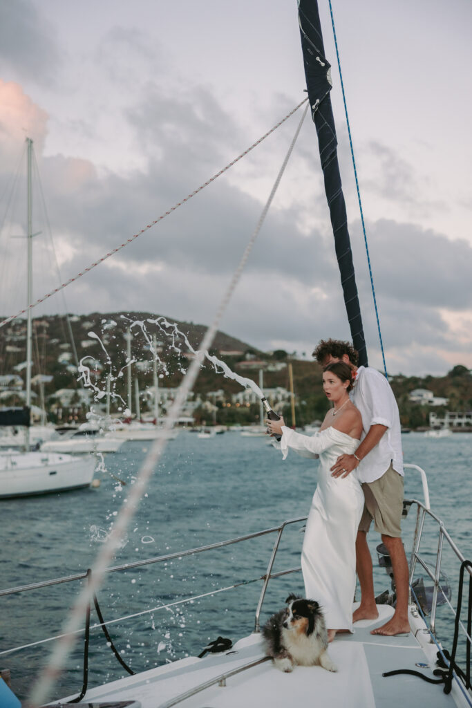 Couple popping a bottle of champagne on their sailboat with their dog at their feet after their st john virgin islands wedding